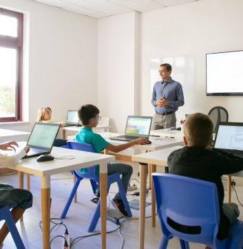 Confident teacher explaining lesson to pupils. Multiethnic children sitting at table in classroom, listening middle-aged man and using laptop computers. Childhood and digital education concept